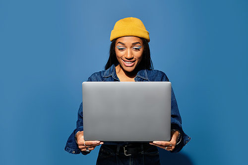 A young woman in a stylish outfit smiles at her laptop in a cheerful setting.