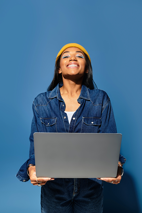 Smiling young woman in a cozy outfit stands with a laptop against a blue background