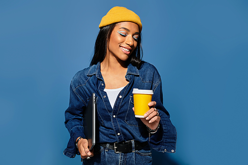 A stylish young woman in a yellow beanie enjoys a warm drink with a joyful smile.