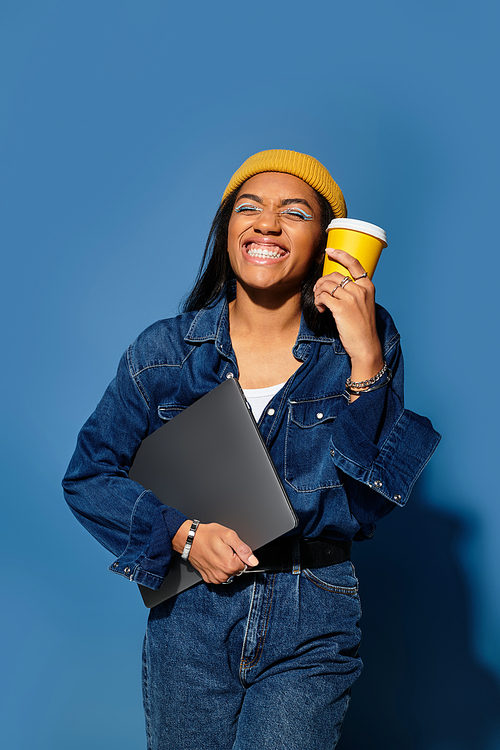 A cheerful young woman in warm autumn clothing holds a coffee cup and laptop, exuding joy.