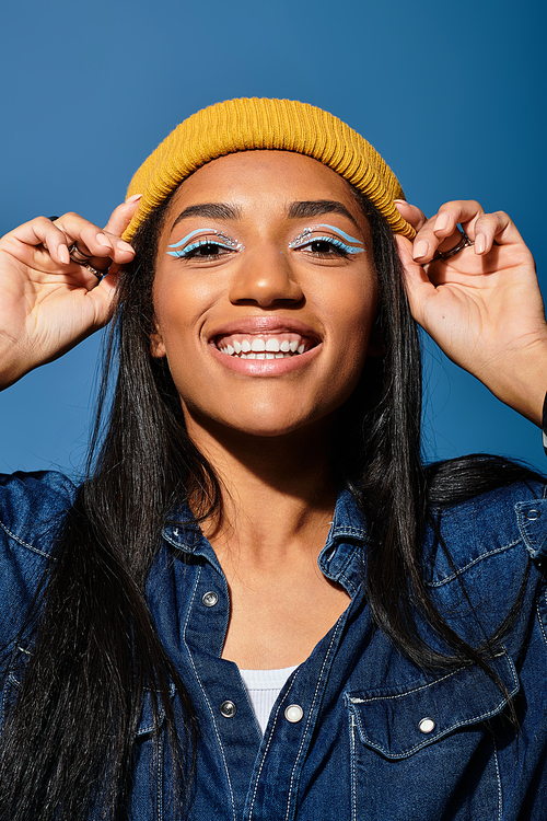 A cheerful young woman showcases her autumn fashion with a yellow hat against a blue backdrop.