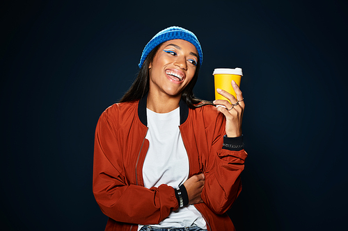 A young woman wears trendy fall attire and holds a yellow cup, exuding joy in a cozy atmosphere.