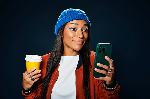 A stylish young woman smiles as she holds her drink and looks at her phone outdoors in autumn.