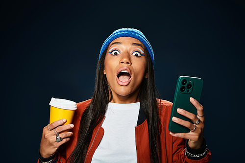 A young woman in warm autumn clothing joyfully reacts while holding coffee and a smartphone.