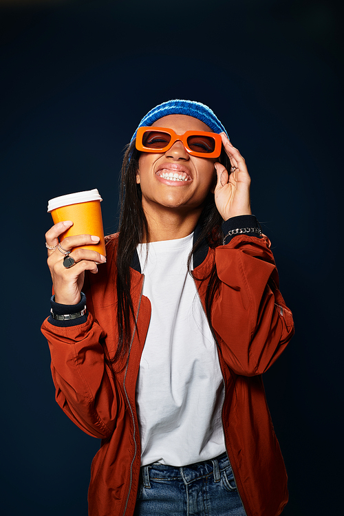 The young woman, wearing orange sunglasses, smiles brightly while holding a warm drink.