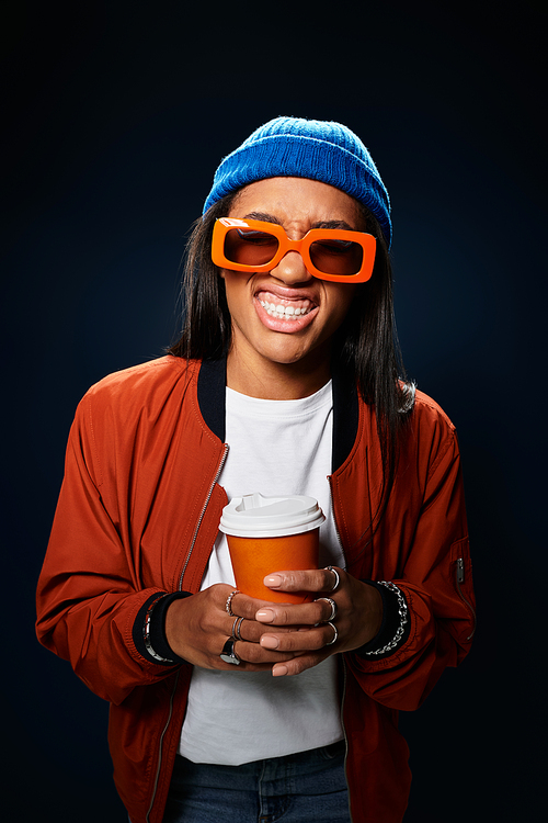 Stylish young woman smiles brightly holding her coffee cup in cozy autumn outfit and blue beanie.