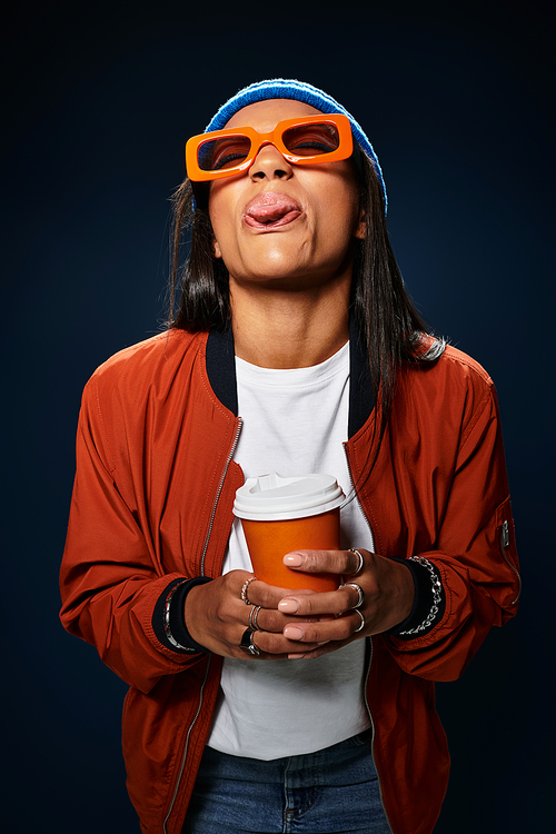 A young woman in cozy autumn fashion enjoys a hot drink while playfully sticking out her tongue.