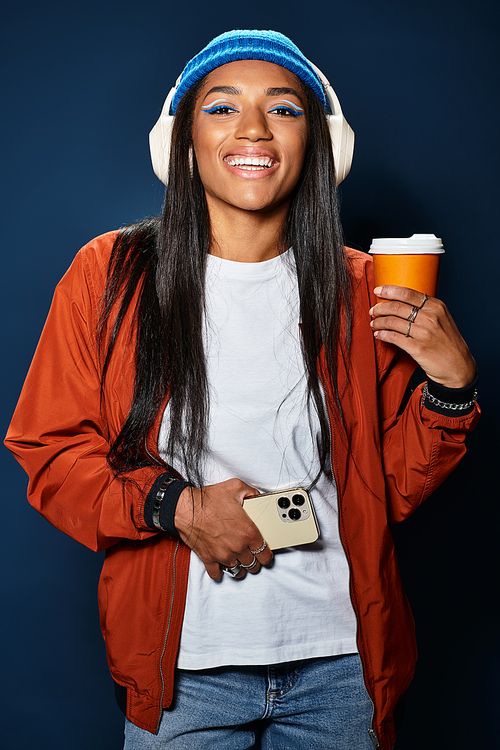 She smiles cheerfully while holding a coffee cup, dressed in warm autumn attire.