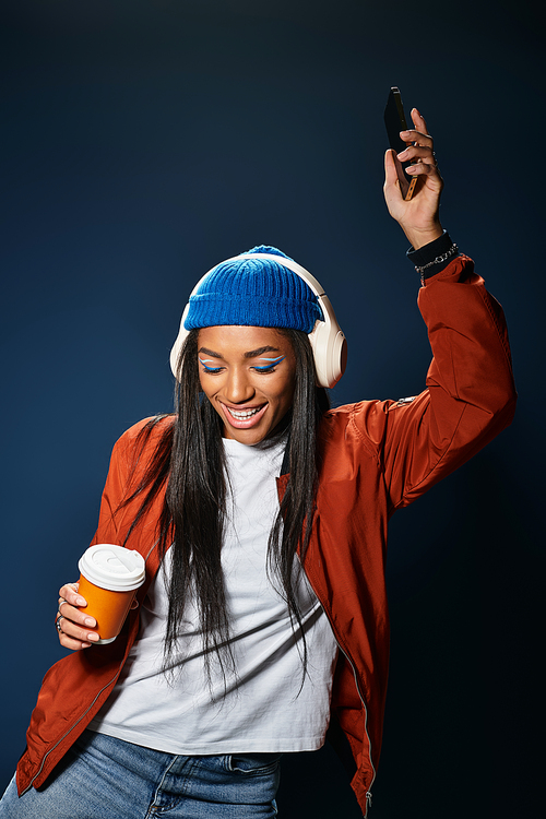 A stylish young woman with headphones dances joyfully while holding coffee in a cozy autumn look.