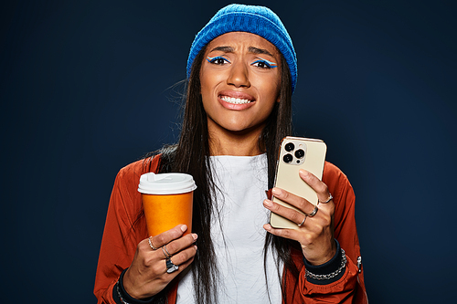 A young woman dressed in autumn fashion expresses mixed emotions while holding a drink and phone.