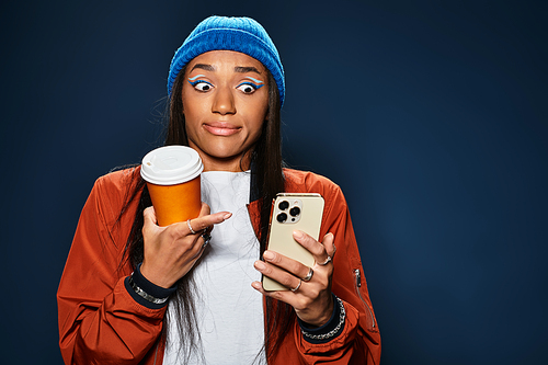 A stylish young woman in warm autumn clothing holds coffee and looks surprised at her phone.