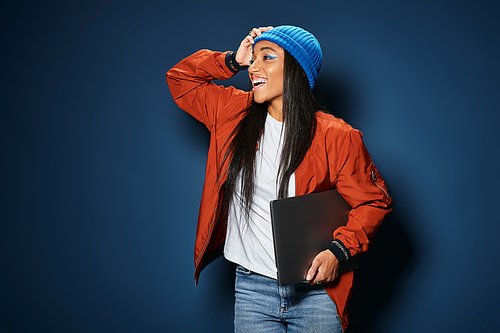 A joyful young woman in warm autumn fashion smiles and holds a laptop under her arm.