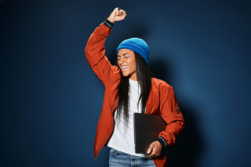 A young woman in a stylish orange jacket and blue beanie raises her fist in exhilaration.