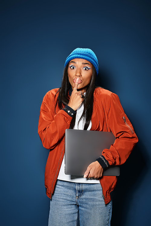 In a stylish warm outfit, a young woman playfully gestures for quiet while holding a laptop.