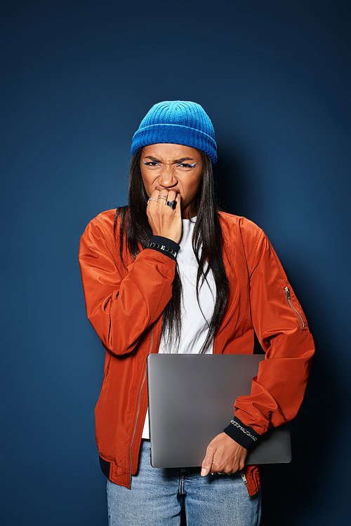 A stylish young woman wearing a cozy jacket and beanie looks worried while gripping her laptop.