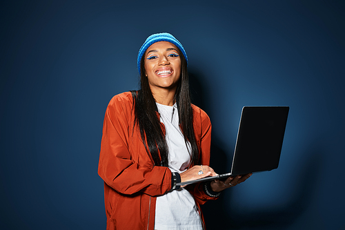 A cheerful young woman dressed in autumn layers joyfully works on her laptop indoors.