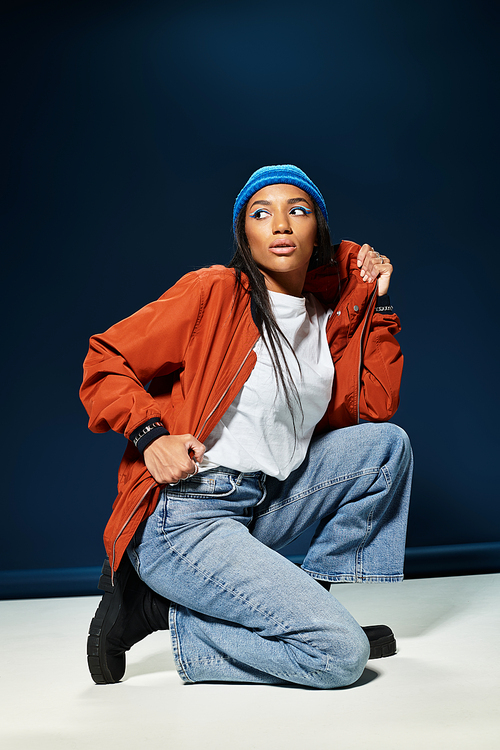 A fashionable young woman showcases her autumn style while kneeling gracefully in a studio setting.