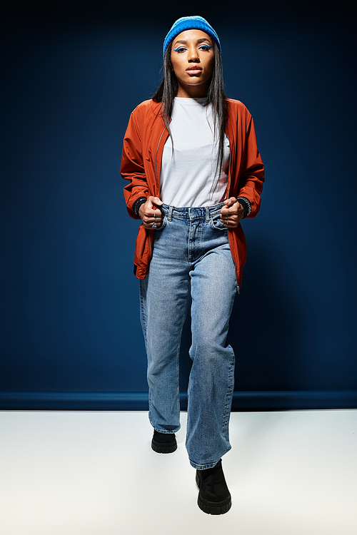 A young woman strolls confidently, flaunting stylish autumn attire against a deep blue backdrop.