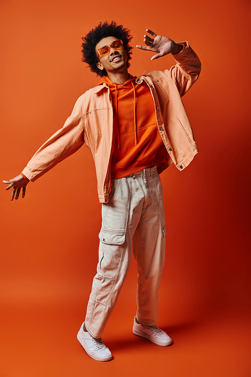 A trendy young African American man with curly hair poses confidently in shirt and white pants against a vibrant orange background.