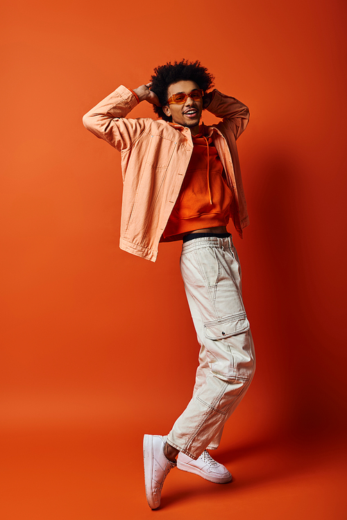 A fashionable young African American man with curly hair and sunglasses posing confidently in front of a bright orange backdrop.