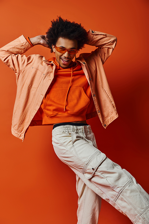 Stylish young African American man in orange shirt and white pants, wearing sunglasses, exuding confidence on orange background.