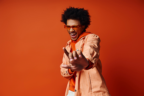 A stylish African American man with curly hair wearing an orange shirt and white pants, showcasing trendy fashion on an orange background.