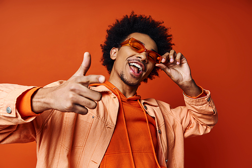 A stylish young African American man in an orange shirt and sunglasses making a silly face on an orange background.