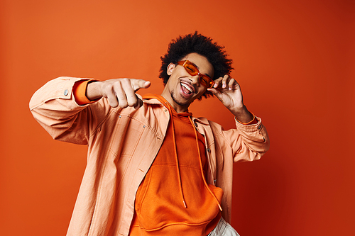 A stylish young African American man with curly hair wearing an orange shirt and sunglasses, posing energetically against an orange background.