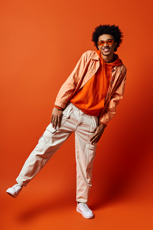 A stylish young African American man with curly hair wearing an orange shirt and khaki pants, sporting sunglasses on an orange background.