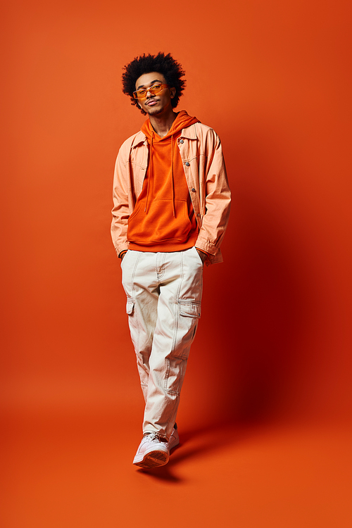 Stylish young African American man in trendy attire and sunglasses exuding emotion in front of a striking orange backdrop.