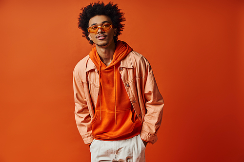 A curly African American man exuding style and emotion in trendy orange shirt, white shorts, and sunglasses on an orange background.