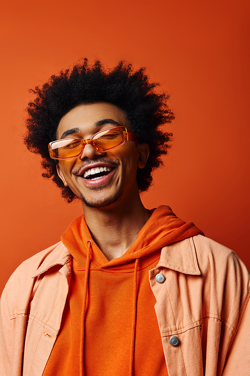 Stylish young African American man in orange hoodie and sunglasses with curly afro hair on vibrant orange background, expressing emotions.