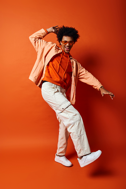 A trendy African American man with curly hair and sunglasses, dressed in an orange shirt and white pants, poses against an orange background.