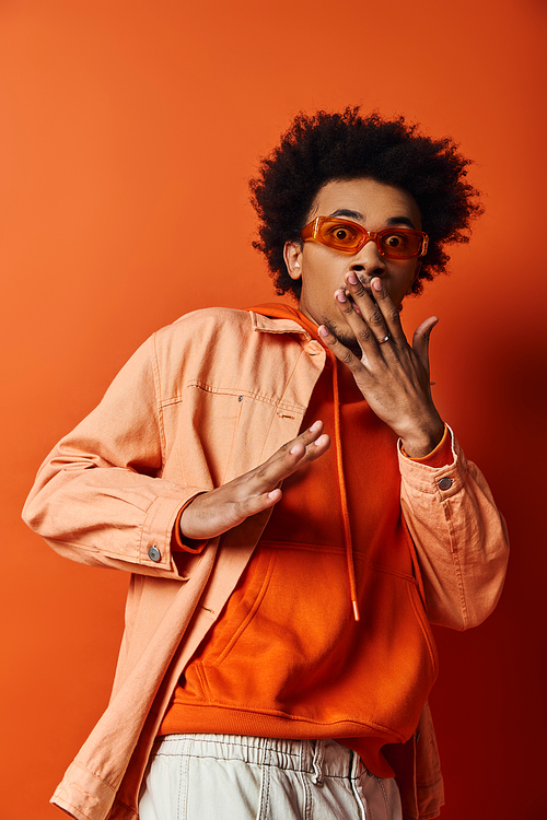 A trendy young African American man in an orange shirt and sunglasses holds his hands up to his mouth in a gesture of emotion.