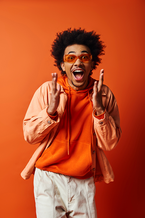 A stylish young African American man in an orange shirt and white shorts exudes emotions against an orange background.