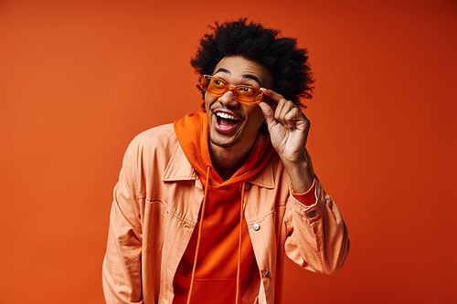 A young African American man with curly hair in trendy attire and sunglasses, showcasing emotions against an orange background.