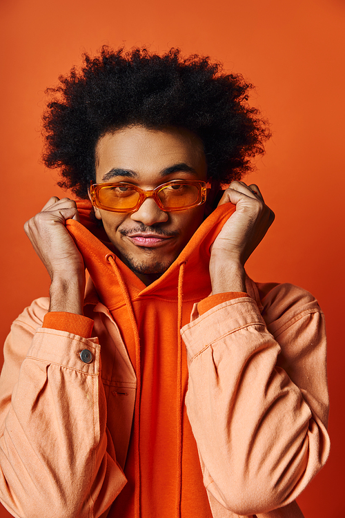 A stylish young African American man with curly hair wearing an orange jacket and trendy sunglasses against a vibrant orange background.