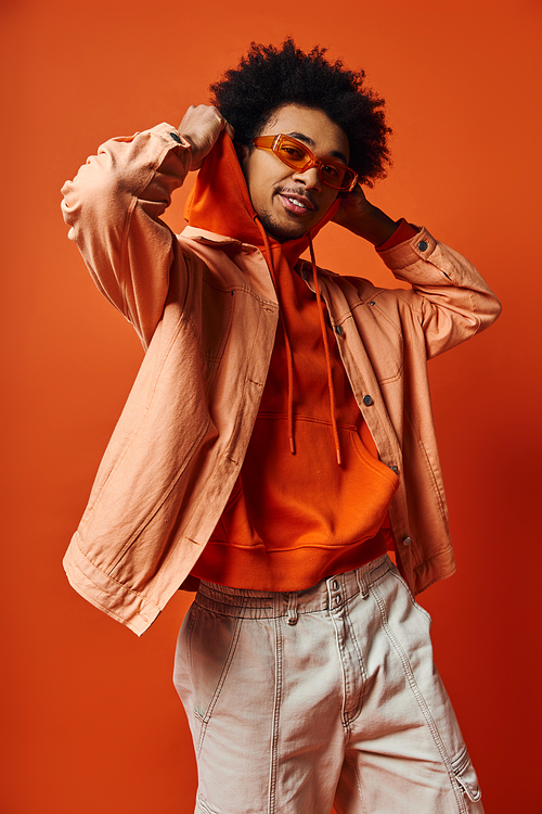 A stylish young African American man with curly hair wearing an orange shirt and white shorts, exuding confidence on an orange background.