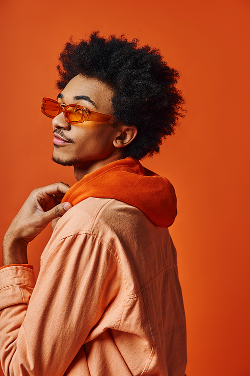 A stylish young African American man with curly hair wears sunglasses and a trendy attire on an orange background.