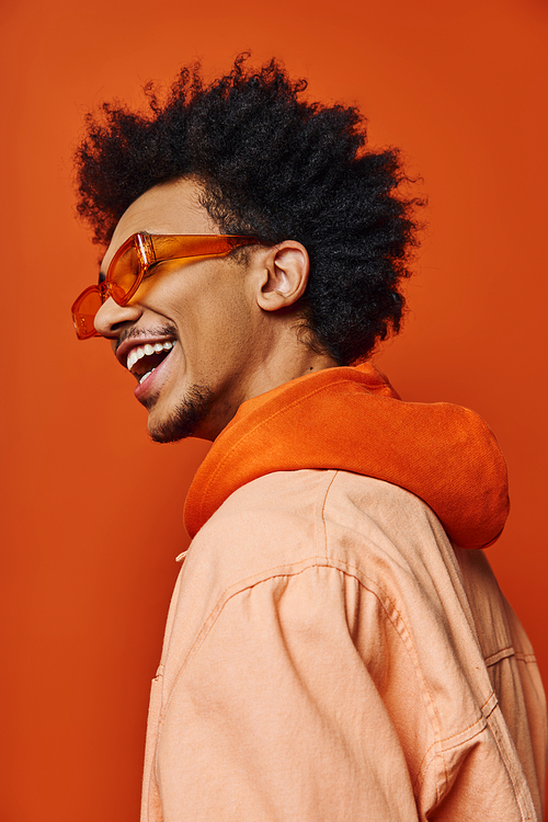 A stylish young African American man with a curly afro wearing sunglasses and a jacket, expressing emotions on an orange background.