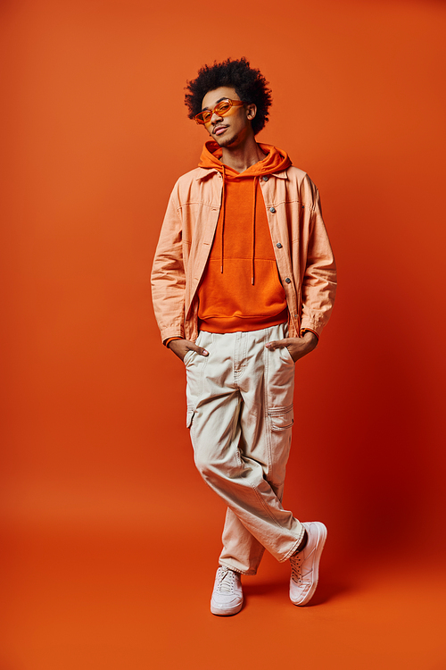 Trendy African American man with curly hair, wearing sunglasses, posing emotionally in bright orange hoodie and khaki pants.