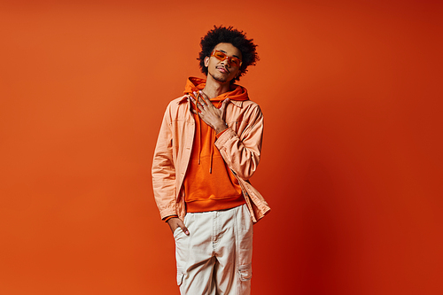 A trendy, curly African American man poses in an orange jacket and white pants against a bold orange backdrop, exuding confidence and style.