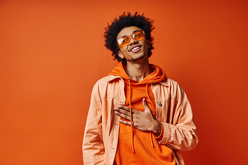 A curly African American man exudes emotion in trendy orange hoodie and sunglasses on bright orange backdrop.