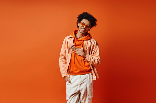 Stylish young African American man with curly hair wearing trendy orange jacket, white pants, and sunglasses on vibrant background.