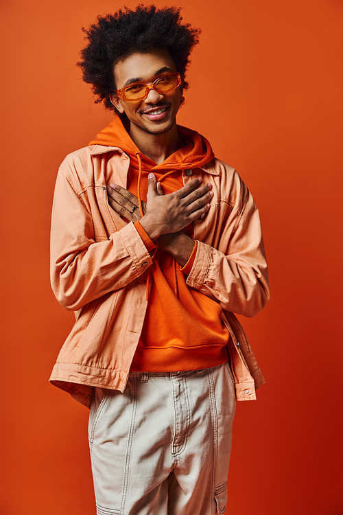 A young African American man with curly hair wearing an orange jacket and sunglasses stands confidently against a bright orange background.