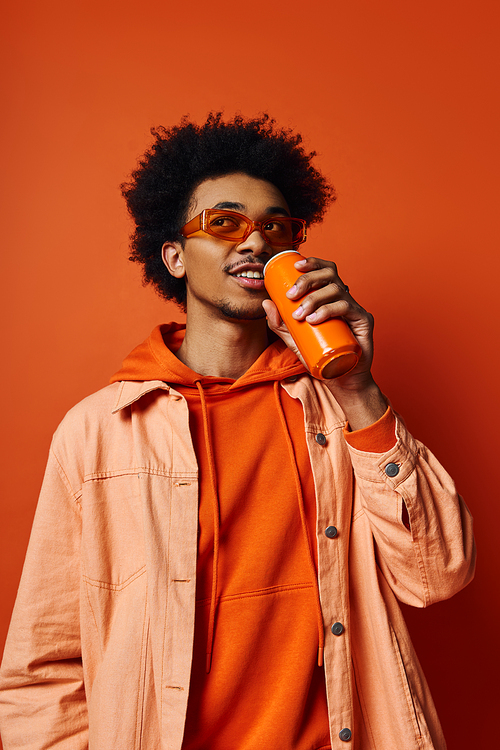 A trendy African American man, curly hair, orange hoodie, drinks from a can, sunglasses, emotional expression on orange background.