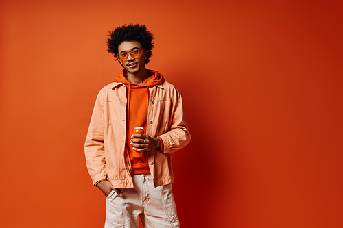 Stylish young African American man posing confidently in trendy orange jacket and white pants on a vibrant orange backdrop.