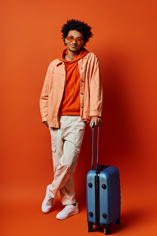 A young African American man stands next to a blue suitcase, exuding style and emotion against an orange backdrop.