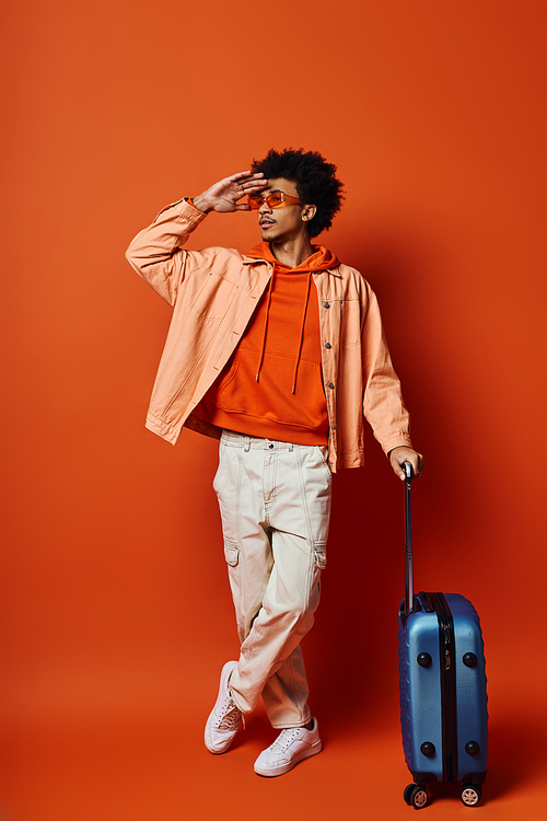 Stylish African American man with curly hair and sunglasses standing next to a blue suitcase on an orange background.