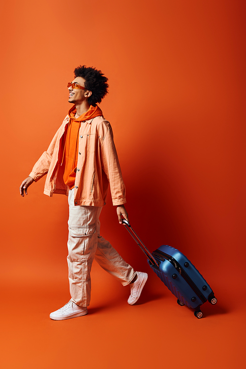 A stylish young African American man with curly hair and trendy attire walking forward while holding a suitcase in his hand against an orange background.